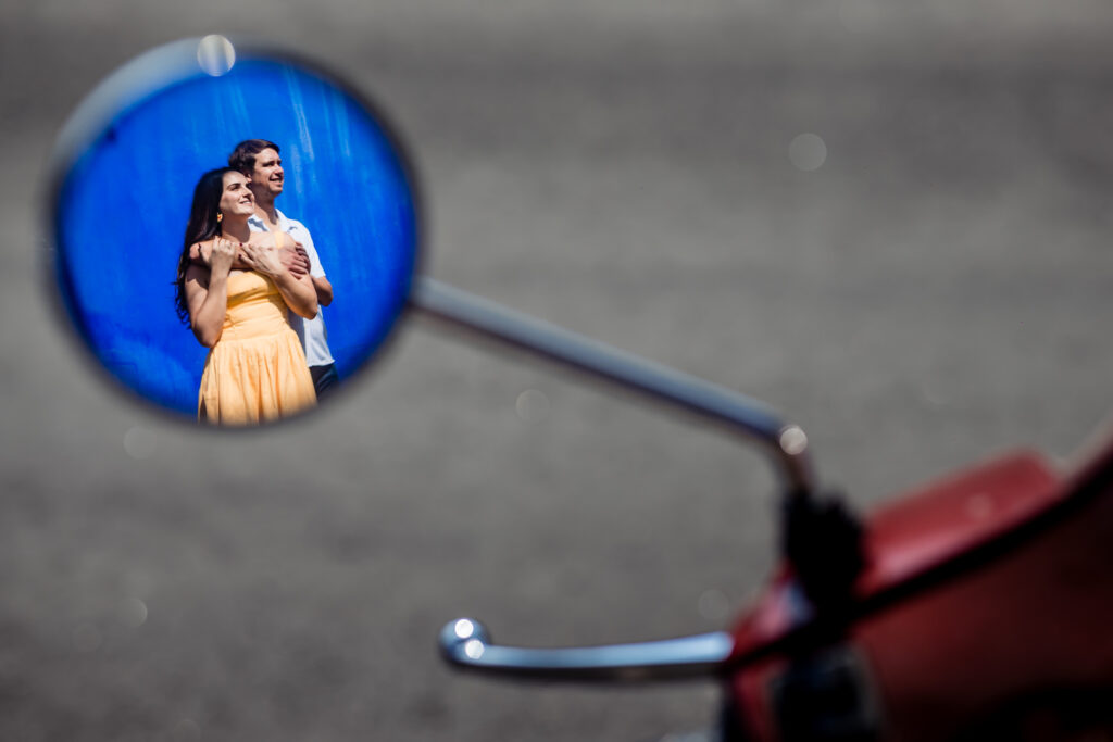 couple hugging each other supported on a blue wall and they are framed in a motor bike's mirror