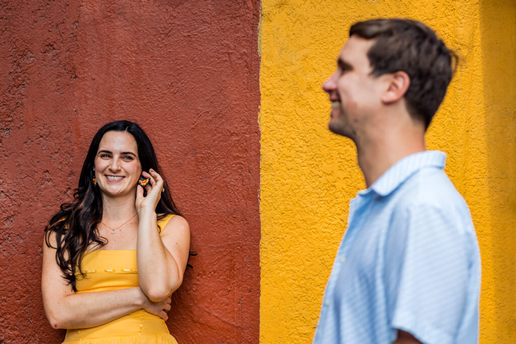 a girl is smiling on the background supported on the wall, on the foreground her boyfriend is smiling too, the wall is divided in two colors red and yellow 
