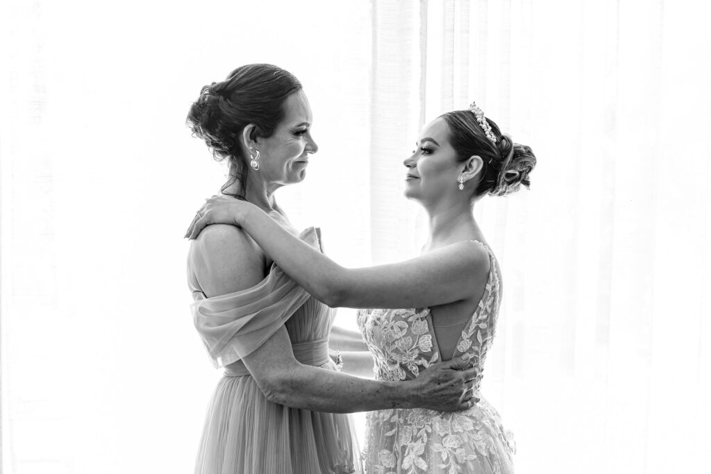 Mum and bride are hug to each other and looking at each other in front of a window, mum is looking her daughter with proud