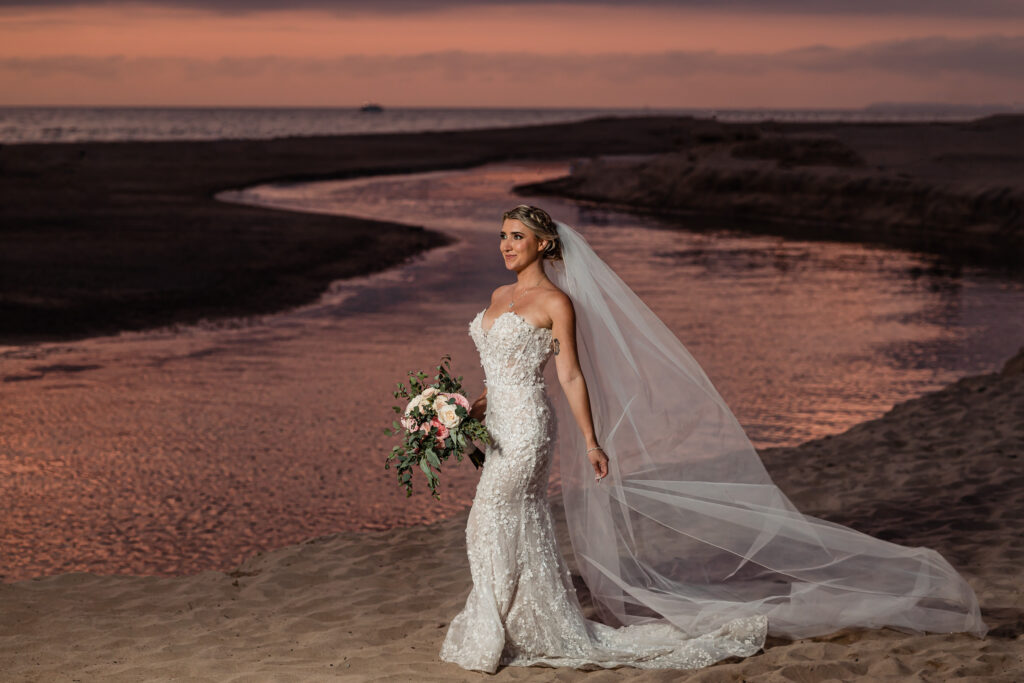 a bride is on the beach with an orange sunset on the background, she have a big veil moving with the wind and she is holding the bouquet