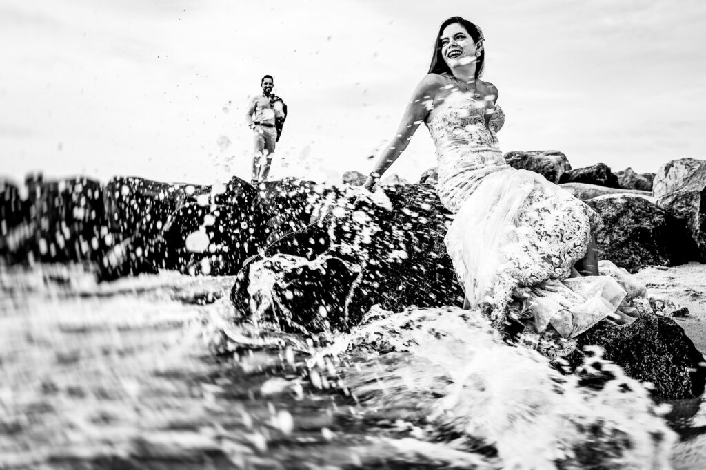 a bride is sit on a rock in the beach of Puerto Vallarta, the groom is on the background holding the suite, a wave is crushing on the rocks