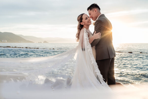 bride and groom hug each other with the ocean on the background, the bride is smiling and the wind is moving the veil in front of the camera