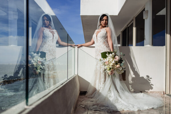 a bride in a white beautiful dress with a bouquet in her hands is on a balcony with the ocean by her side, the glass of the balcony is reflected the bride too