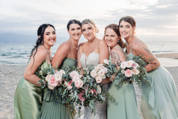 bridemades are hugging the bride on the beach, they are holding flowers and they have the ocean on the background