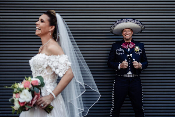bride are in foreground and smiling with her bouquet, on the background the groom is dress as a Mexican charro and smiling