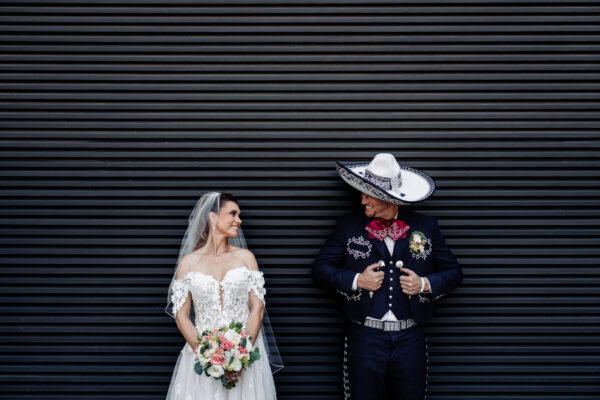 bride and groom are looking at each other and relaxing on a black wall, the groom is dress as a Mexican charro with a big heat