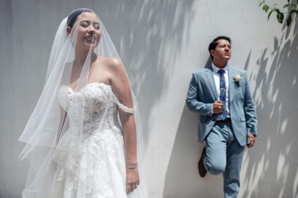 a bride is in the foreground, smiling with the veil on her face on the background there are the groom holding is jacket