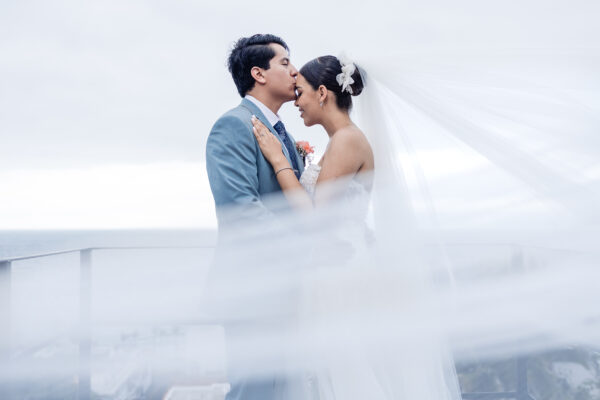 bride and groom are hugging each other and the groom kiss the bride on the forehead, the veil of the wedding dress is in front of the camera