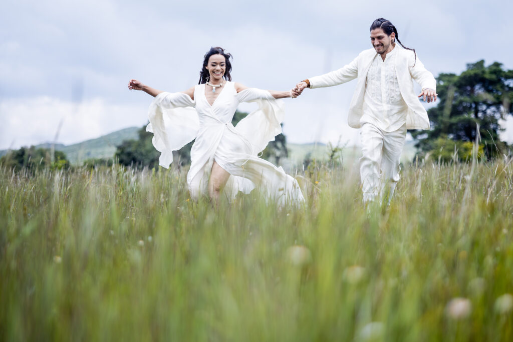 bride and groom are holding hands and running together on a green field, both are smiling