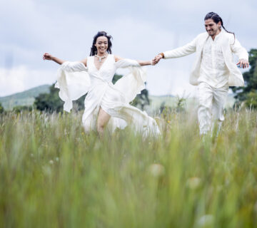 bride and groom are holding hands and running together on a green field, both are smiling