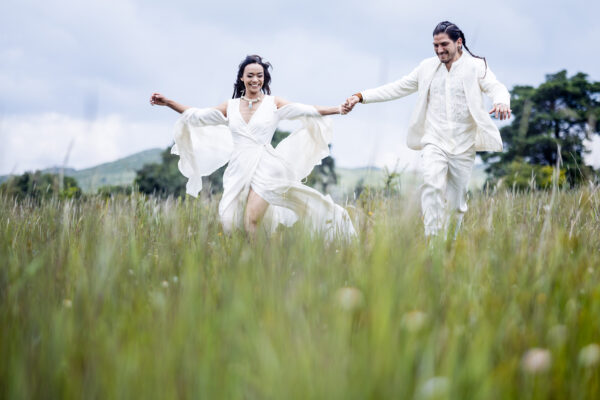 bride and groom are holding hands and running together on a green field, both are smiling