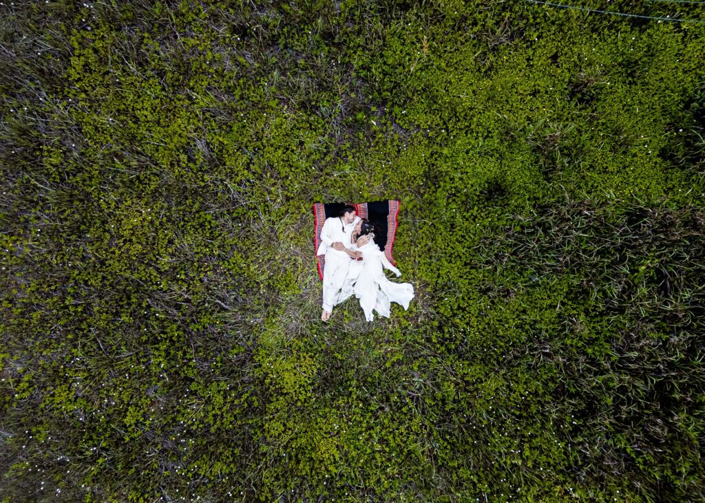 drone shoot from above with the bride and the groom laid down on a green field , both are dressing on white