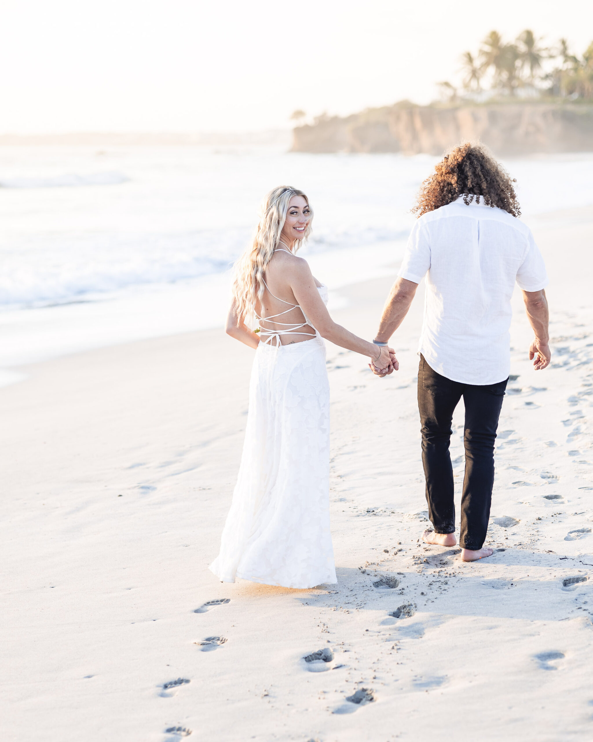 Wedding-bride-groom-love-mita-ocean-8 bride and groom are walking on the beach, the bride is looking to the camera and on the back ground there is a warm light