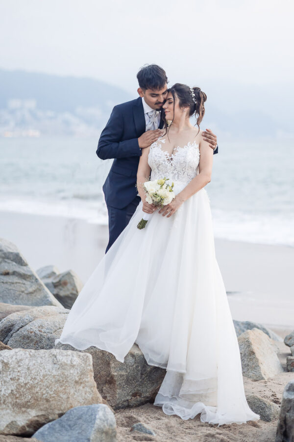 groom is hugging the bride from the back, she il holding the bouquet with both hands, both are on the beach with Puerto vallarta in the background